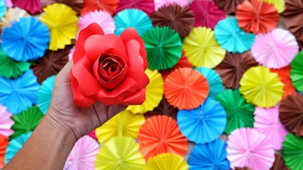 Fototapeta na wymiar Man hand holding red artificial rose paper flower with blur background of many colorful folding circle papers on the floor in art and lifestyle concept