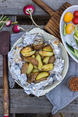Baked potato in foil on skillet with fresh vegetables. Vegan lunch or vegetarian healthy food dinner on wooden table. Top above view.
