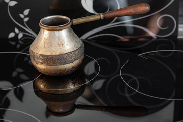 Old Bronze Turkish retro coffee maker kanaka on glass hob and stove with wooden handle, worn on a black background in an apartment in the kitchen. Retro cezve, briki, rakweh, pot on a stovetop.
