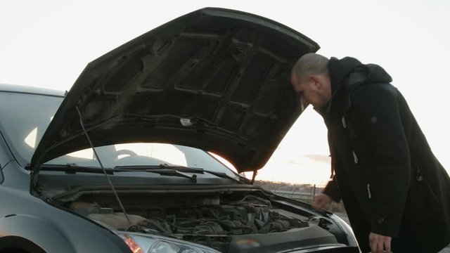 The average plan of a young Caucasian guy with short hair and a Smoking cigarette in his hand, standing near the car black with the hood up. The driver eliminates the damage in the way and scratching