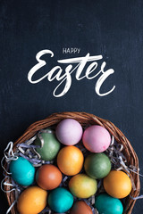 Happy Easter hand lettering inscription and colorful easter eggs in basket on black wooden background.