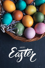 Happy Easter hand lettering inscription and colorful easter eggs in basket on black wooden background.