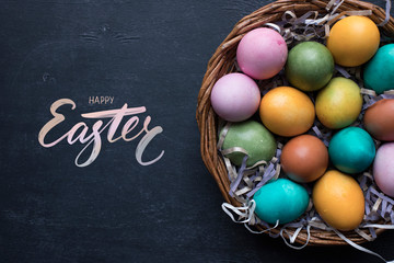 Happy Easter hand lettering inscription and colorful easter eggs in basket on black wooden background.