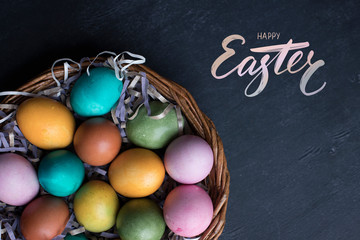 Happy Easter hand lettering inscription and colorful easter eggs in basket on black wooden background.