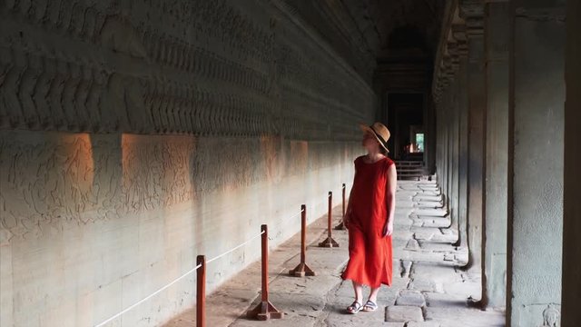Woman in red dress is walking along inner corrdior of Angkor Wat temple and looking at ancient bas-reliefs on the wall. Cambodia