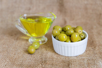  glass sauceboat with extra virgin olive oil and fresh green olives in white ceramic bowl on  burlap cloth background
