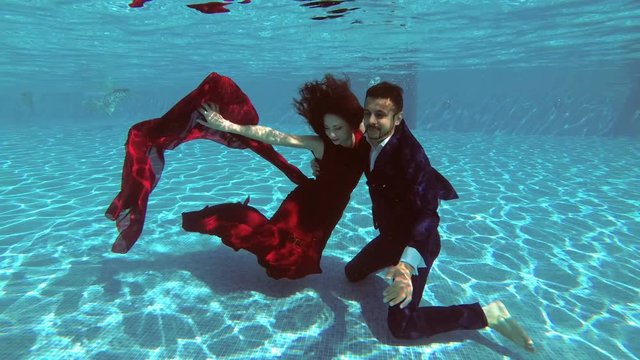 Happy bride and groom sit under the water at the bottom of the pool, smiling and looking at the photographer who takes pictures of them. Slow motion. Underwater photography. 4K. 25 fps