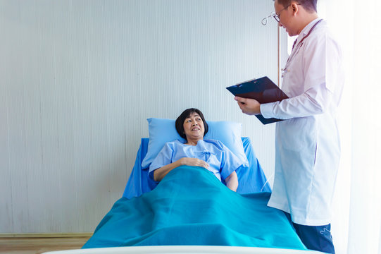 Man Doctor Checking Pressure Of Female Patient, Older Sick Woman Patient Lay On Bed In Hospital With Intravenous Normal Saline.