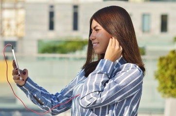 A young girl student of Asian appearance dressed in a striped shirt takes a selfie and listens to...