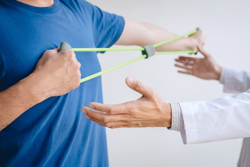 Doctor physiotherapist assisting a male patient while giving exercising treatment on stretching his arm with exercise band in the clinic, Rehabilitation physiotherapy concept