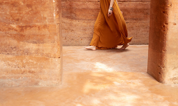 Unrecognisable barefooted woman in a dress walking by the rammed earth walls.