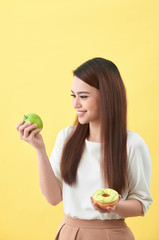 Portrait of a smiling young asian woman choosing between donut and green apple isolated over yellow background