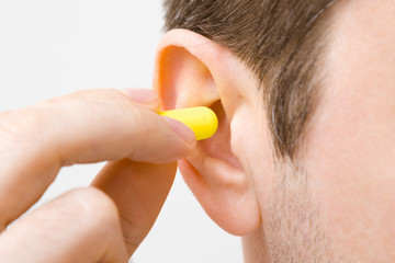 Young man's fingers putting yellow earplug into his ear on light gray background. Closeup. 