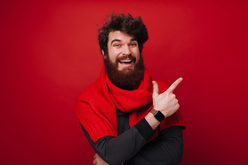 Cheerful bearded man with red scarf pointing away over red wall background