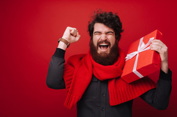 Portrit of excited bearded man, holding a gift box, while celebrating over red background