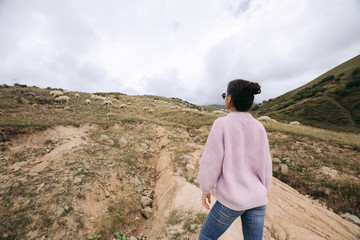 Naklejka premium Happy young woman in a sweater against herds of sheep in the mountains
