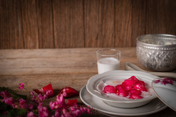 Thapthim krop, mock pomegranate seeds in coconut and syrup in the white bowl on the wood table there are flower, spoon, coconut milk and water bowl placed around.
