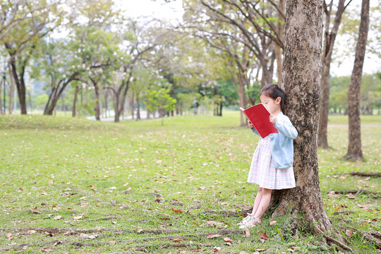Pretty little child girl reading book in park outdoor standing lean against tree trunk in summer garden.
