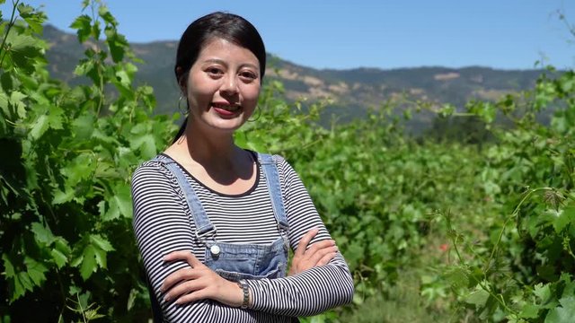 proud young asian woman farmer standing with folded arms in vineyard background. young satisfied female winemaker looking face at camera smiling crossed hands. confident owner of farm outdoor
