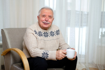 Portrait of Happy and smiling senior male 70-75 years old sitting on the armchair and holding cup of coffee in hand on window background