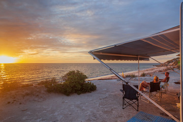 Retired couple sitting under the awning of a of caravan enjoying a wine next the beach with a golden sunset.