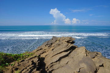 Waves from the ocean to the rock cliffs of the beach on the coast of Bali, with the blue sea and the bright, cloudy sky.