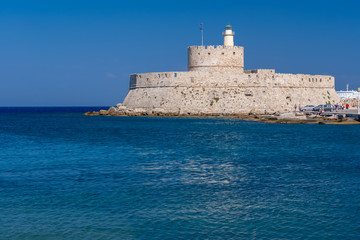 Fototapeta na wymiar Rhodos, Greece - August 2016: Fort of Saint Nicholas at the entrance in the Mandraki old harbour of the City of Rhodes. Stone fortress or castle on the seashore against a clear blue sky and water.