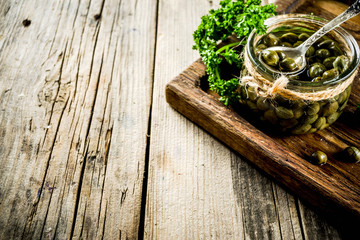 Pickled capers in little jar, with herbs on rustic wooden background copy space