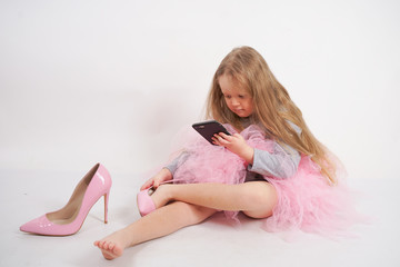 Obraz na płótnie Canvas a little caucasian child girl sits and puts mother's high-heeled pink shoes, taking pictures of it all and selfies on her smartphone on white studio background