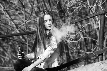 Fototapeta na wymiar Vape teenager. Young pretty white girl in casual clothing smoking an electronic cigarette on the old wooden stairs on the street in the spring. Bad habit. Vaping activity. Black and white.