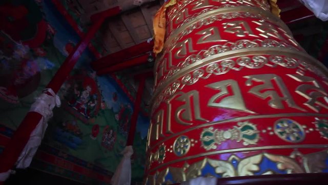 An ornately decorated column spins in a Nepal monastery, covered in golden script, with a wall depicting a colorful mural in background.