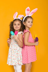 two little girls with Easter bunny ears holding colorful eggs on yellow background. Happy easter. cute children