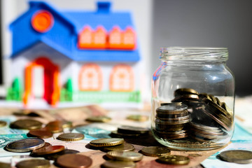 Glass jar with coins and house model on the blurred background. By a new home concept.