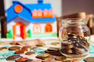 Glass jar with coins on money field and model of house on the blurred background. Saving money to buy a house concept.