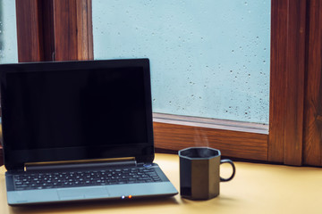 Fototapeta premium Computer and a cup of hot tea against the window with raindrops background. Cosy workplace, freelance. Copy space.