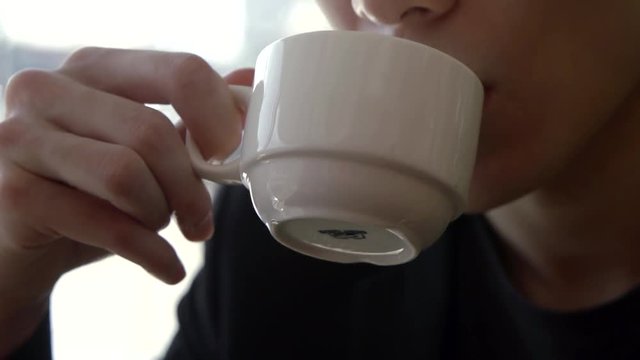 A dark-haired Asian guy is close-up drinking from a Cup of coffee and looking at the camera.