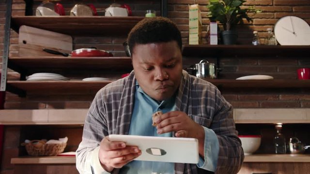 Shot of african American man use digital tablet computer at kitchen eating homemade cookies relaxed american black home technology touchscreen wireless eating morning close up slow motion