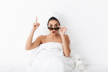 Excited woman with towel on head lies in bed covering body under blanket isolated over white wall background wearing sunglasses have an idea pointing.