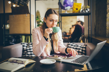 Beautiful young caucasian woman drinking coffee and typing on a keyboard inside a cafe at a wooden table. The topic of modern professions is a blogger, freelancer and writer