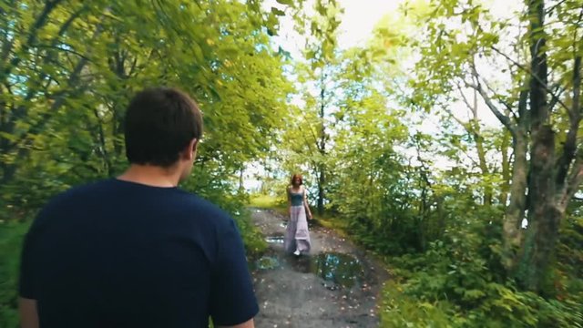 Young man and beautiful young woman walking towards each other on a dirty path with puddles surrounded by green trees in the park. Unexpected meeting.