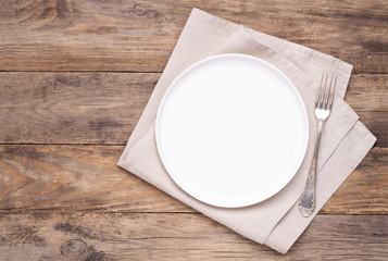 Empty white plate with napkin and silver fork on old wooden table, top view 