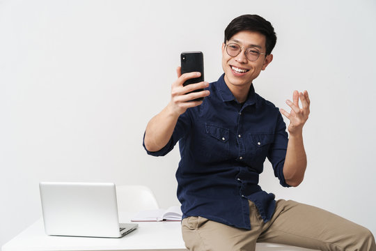 Photo of cheerful asian businessman sitting at table and holding smartphone while working with laptop in office