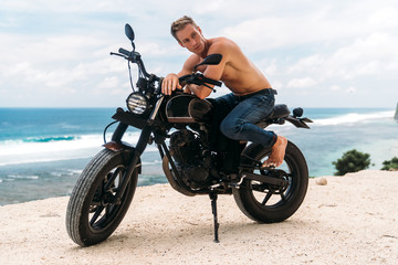 Obraz na płótnie Canvas Sexy athletic man with perfect naked body sitting on motorbike, ocean waves and beautiful mountains on background