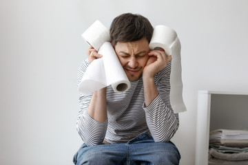 Stressed man with rolls of paper sitting on toilet bowl at home