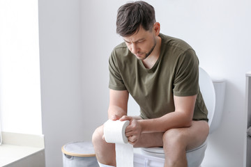 Man with roll of paper sitting on toilet bowl at home