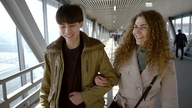 A young couple, an Asian black unbuttoned jacket and a curly-haired girl in a coat, go hand in hand at the station, talk and laugh.