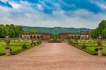 The lovely walkway to the Baroque garden of Weikersheim Palace, framed by two statues, is leading to the beautiful Hercules Fountain in the middle and the picturesque orangery in the background. - obrazy, fototapety, plakaty