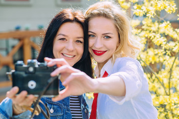 Outdoor portraits of two pretty girls friend taking selfie on vintage camera, enjoy nice day, looking on camera and making funny faces over yelow blossom tree. Travel, lifestyle concept.