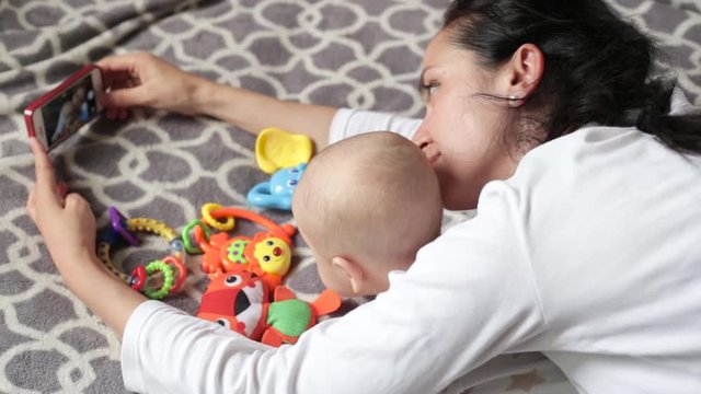 mother with baby makes selfie, lying on the bed. toys are near