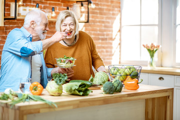 Cheerful senior couple eating salad standing together with healthy food on the kitchen at home....